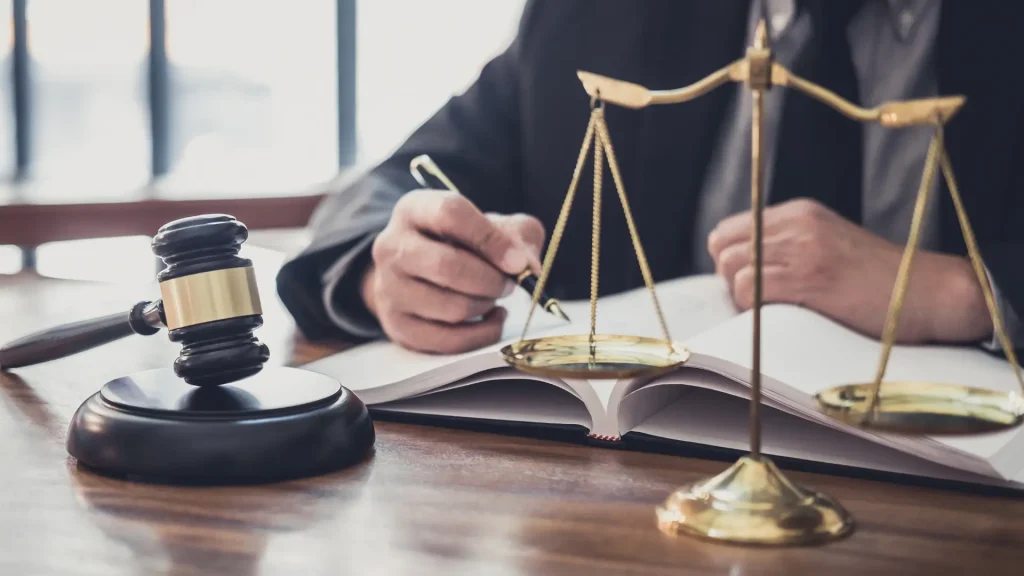 An image of a legal setting, featuring a person writing in a book with a golden balance scale and a gavel on the desk, symbolizing law and justice.
